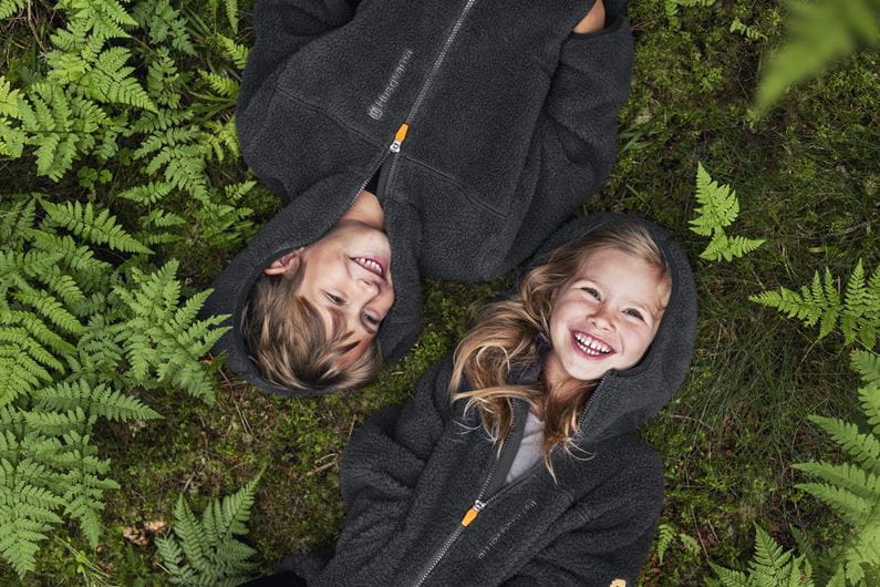Två glada barn ligger på en bädd av gröna ormbunkar, båda klädda i svarta Husqvarna Xplorer Kids luvtröjor med orangea dragkedjedetaljer. De ser upp mot kameran med leende ansikten.