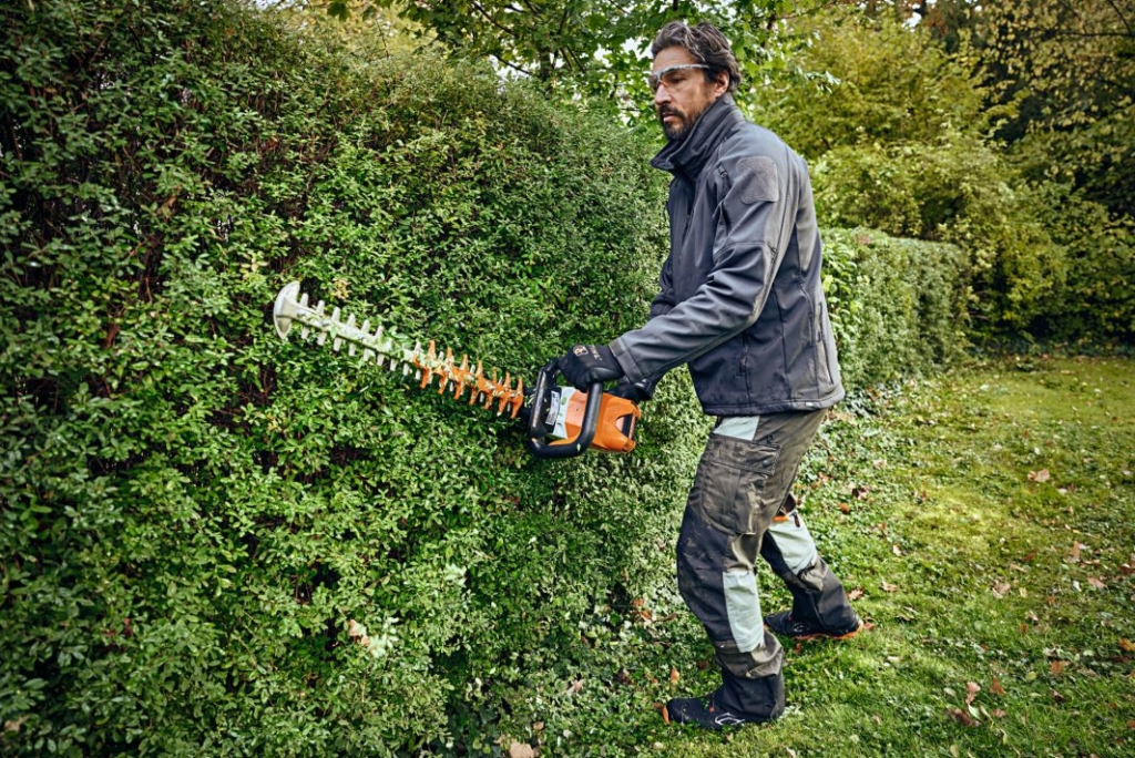 En man i skyddskläder klipper en tät häck med sin Stihl HSA 100 Batterihäcksax i en grönskande trädgård