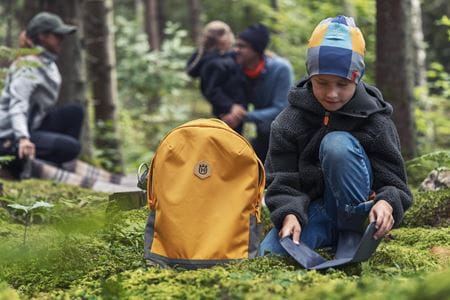 En pojke sitter i skogen iförd en Husqvarna Xplorer Kids mössa och bredvid honom ligger en Husqvarna väska. I bakgrunden ser man andra personer njuta av naturen