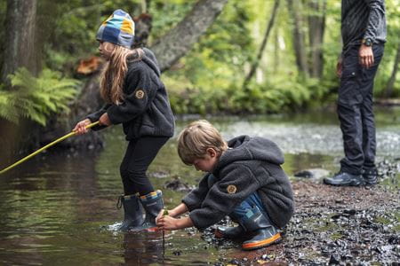 Två barn i skogen, båda klädda i Husqvarna kläder, där en av dem bär en Husqvarna Xplorer Kids mössa. De leker vid en bäck, medan en vuxen står i bakgrunden