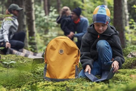 En pojke i skogen bär en Husqvarna Xplorer Kids mössa och sitter bredvid en gul Husqvarna Xplorer Kids ryggsäck. I bakgrunden ser man andra personer njuta av naturen